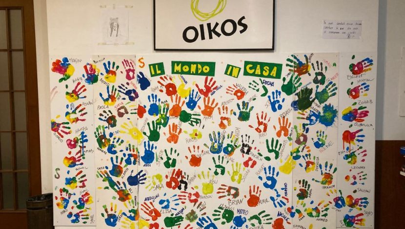 In alto un quadro col logo di Oikos Onlus, in basso, le impronte di tante mani colorate, con dei nomi scritti accanto