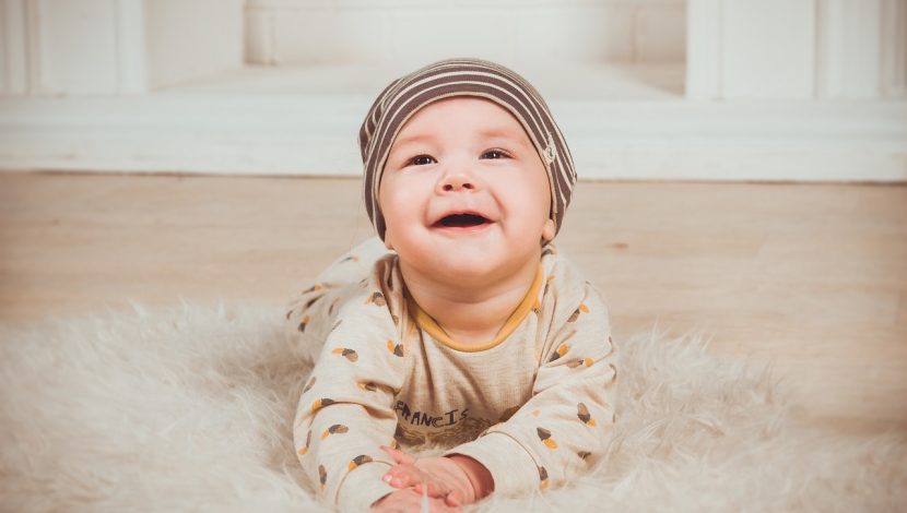 Un bambino di pochi mesi a pancia in giù su un tappeto peloso beige. Guarda in alto sorridendo, ha una cuffietta