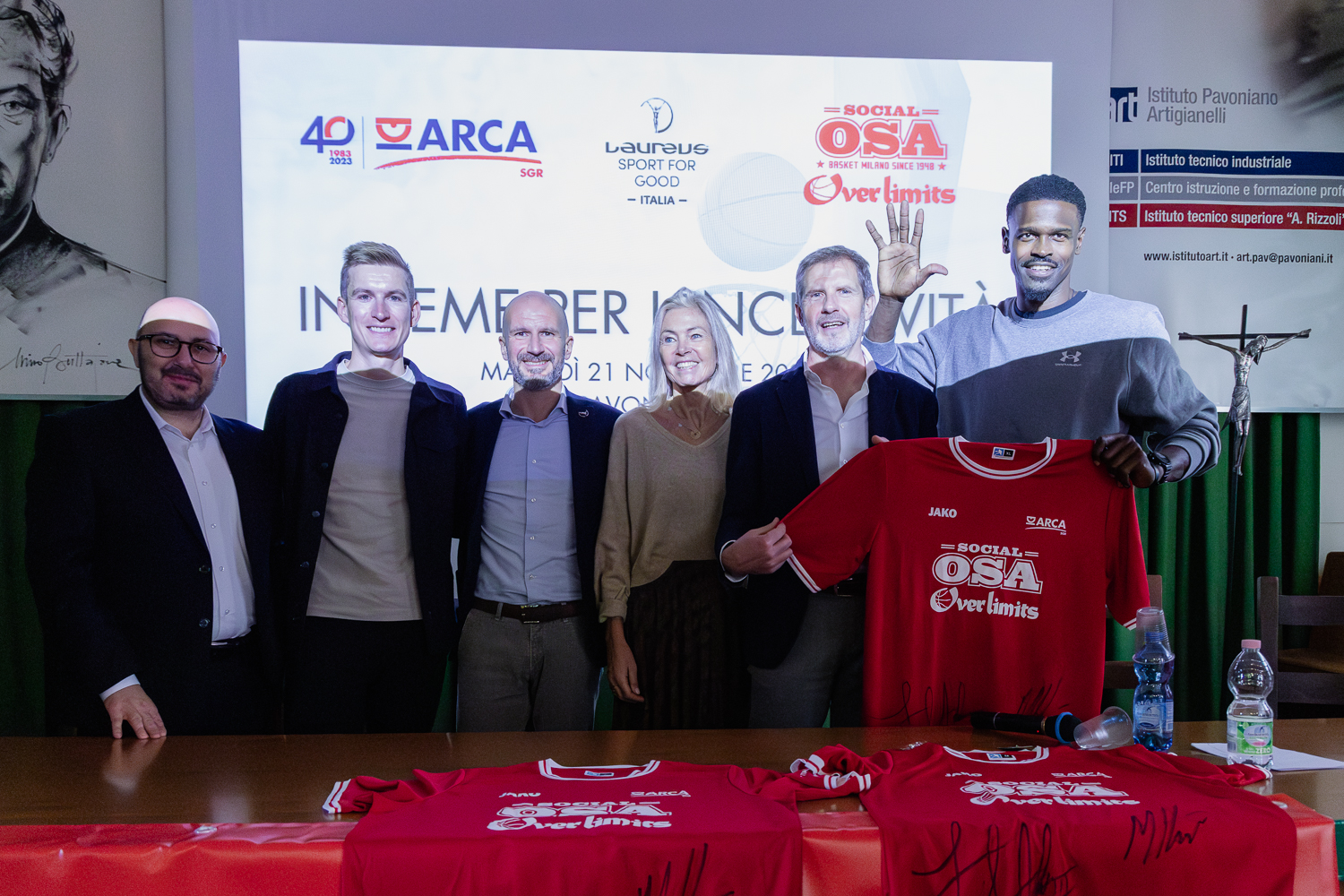 cinque persone sorridenti, davanti a un tavolo con magliette rosse. La persona più a destra mostra una mano aperta 