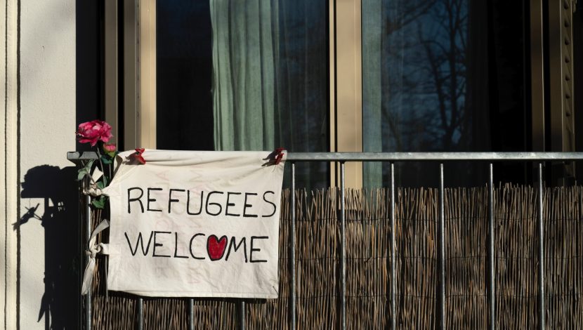 Un balcone con a sinistra appeso alla ringhiera un cartello "refugees welcome", con un cuore al posto della o e una rosa appesa vicino
