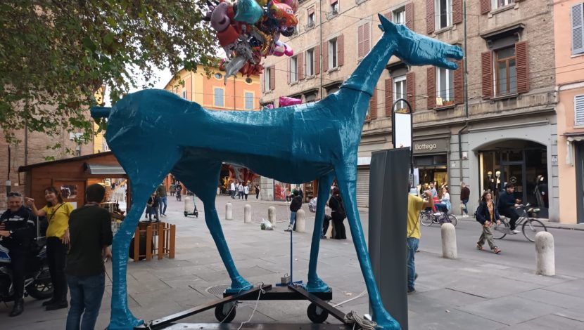Una statua di un cavallo blu in una via cittadina, dietro ci sono dei palloncini