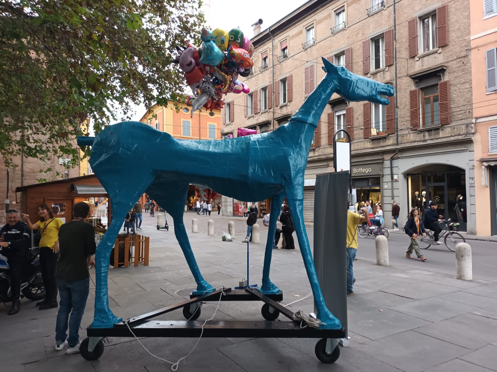 Una statua di un cavallo blu in una via cittadina, dietro ci sono dei palloncini