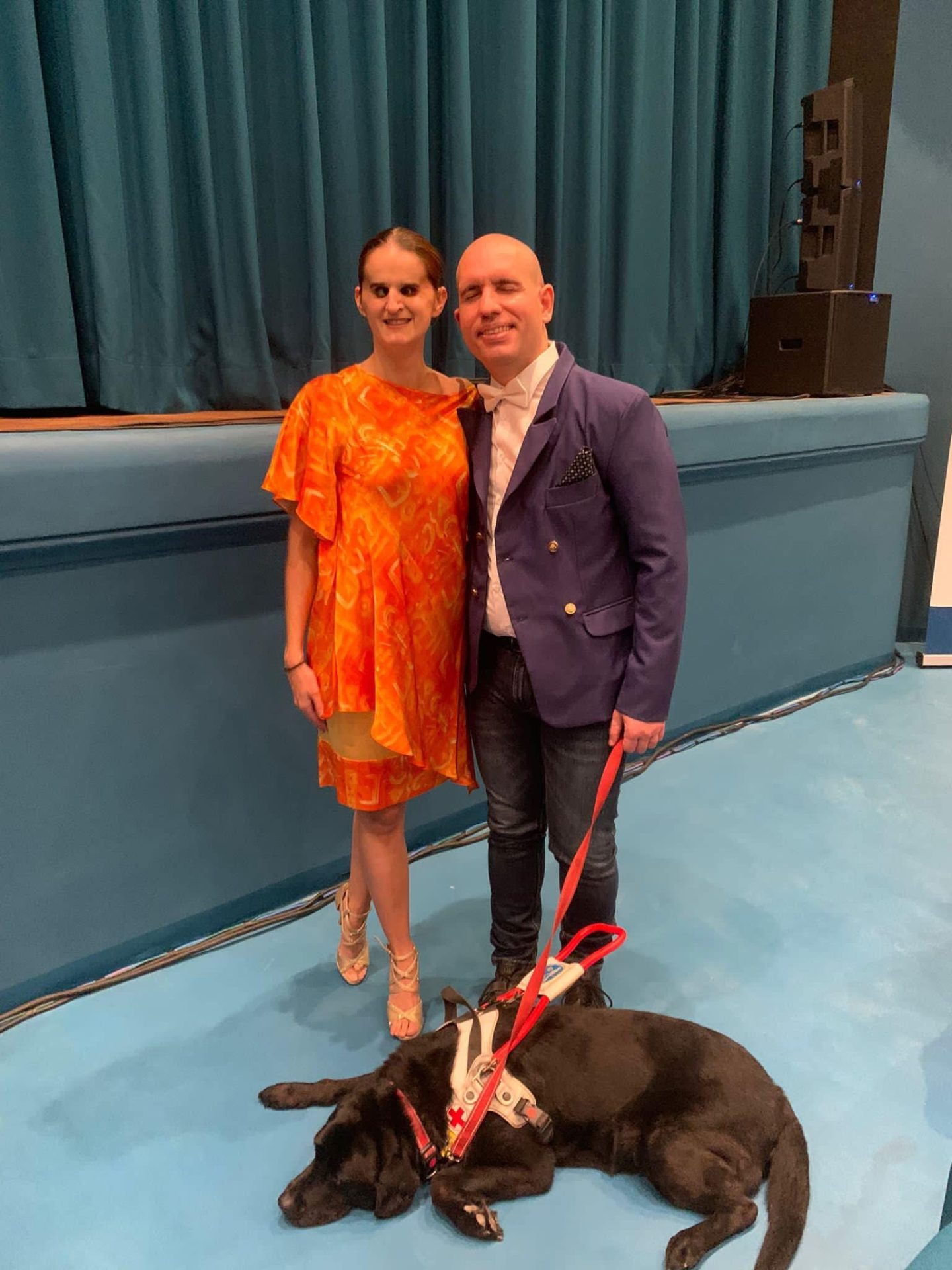 Travaini e Lachin in un teatro, con vestiti eleganti, sotto al palco. Lachin tiene il guinzaglio di un cane guida