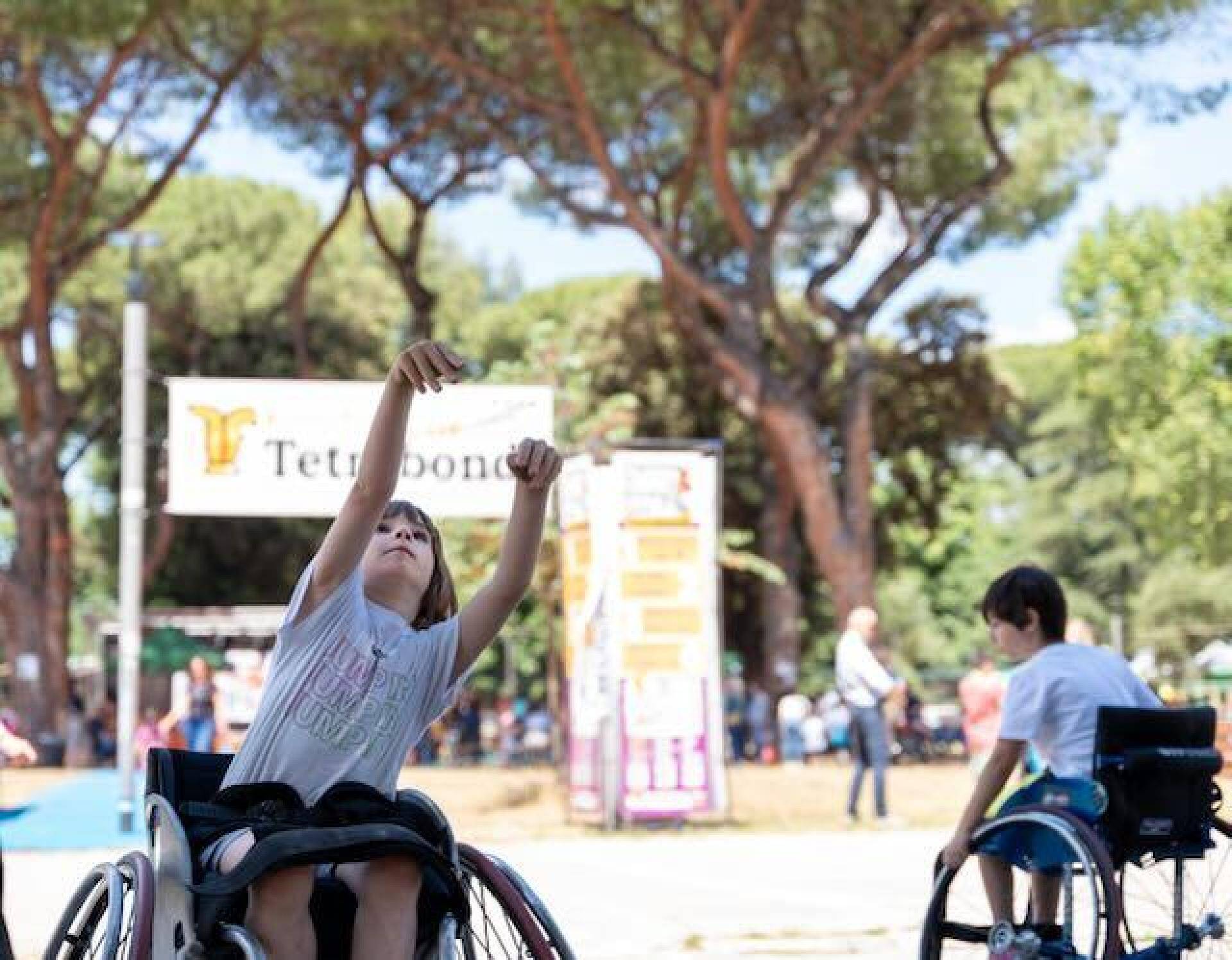 Un bambino in primo piano seduto su una carrozzina nell'atto di lanciare una palle, dietro, sullo sfondo, tra gli alberi, un banner con la scritta "Tetrabondi"